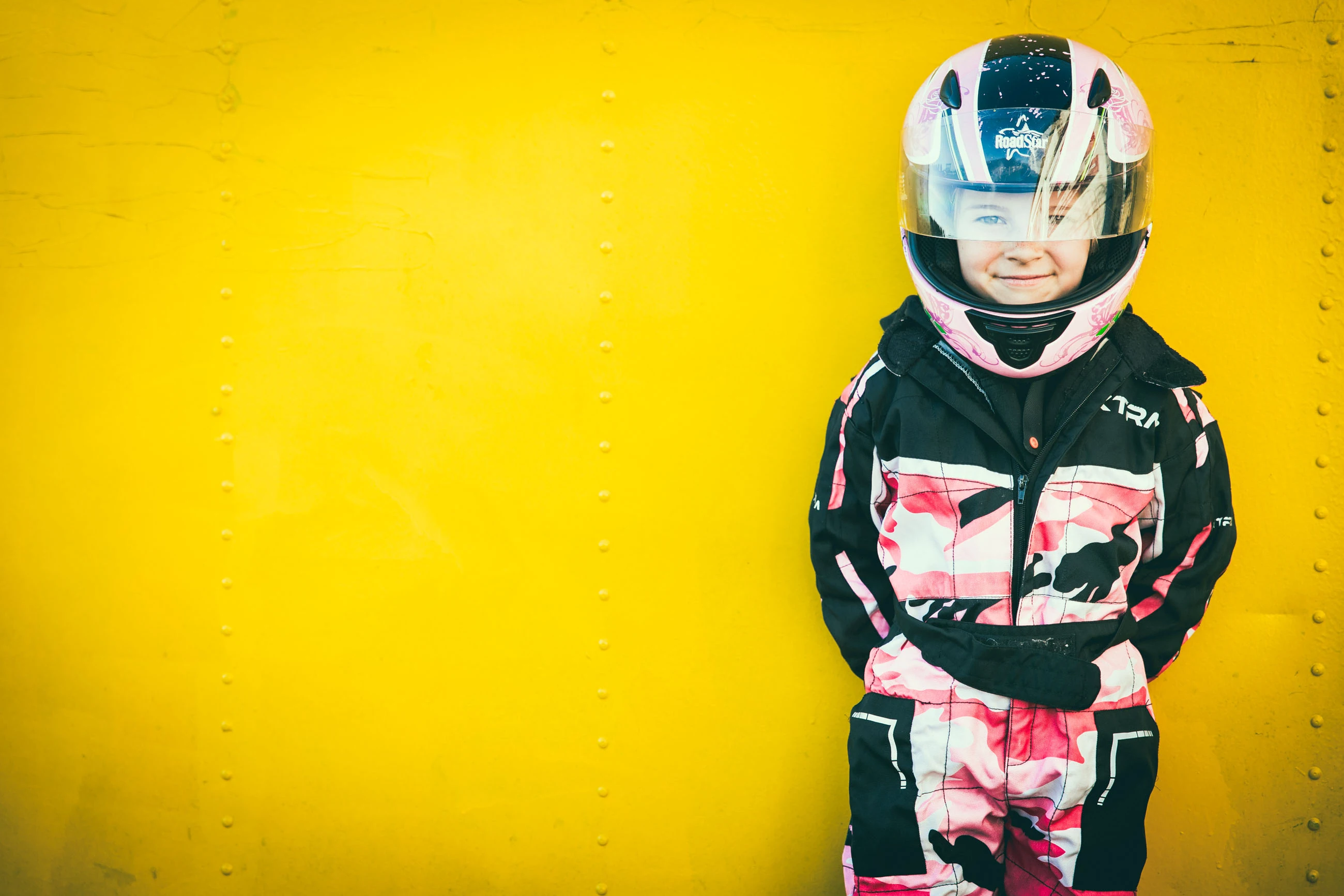 Kind im Kart-Rennanzug mit Helm vor knallgelber Wand