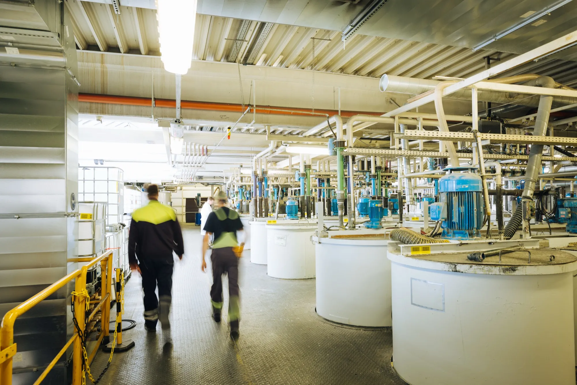 Produktionshalle einer Farbproduktion mit großen Rührbehältern und zwei Mitarbeitern im Gang