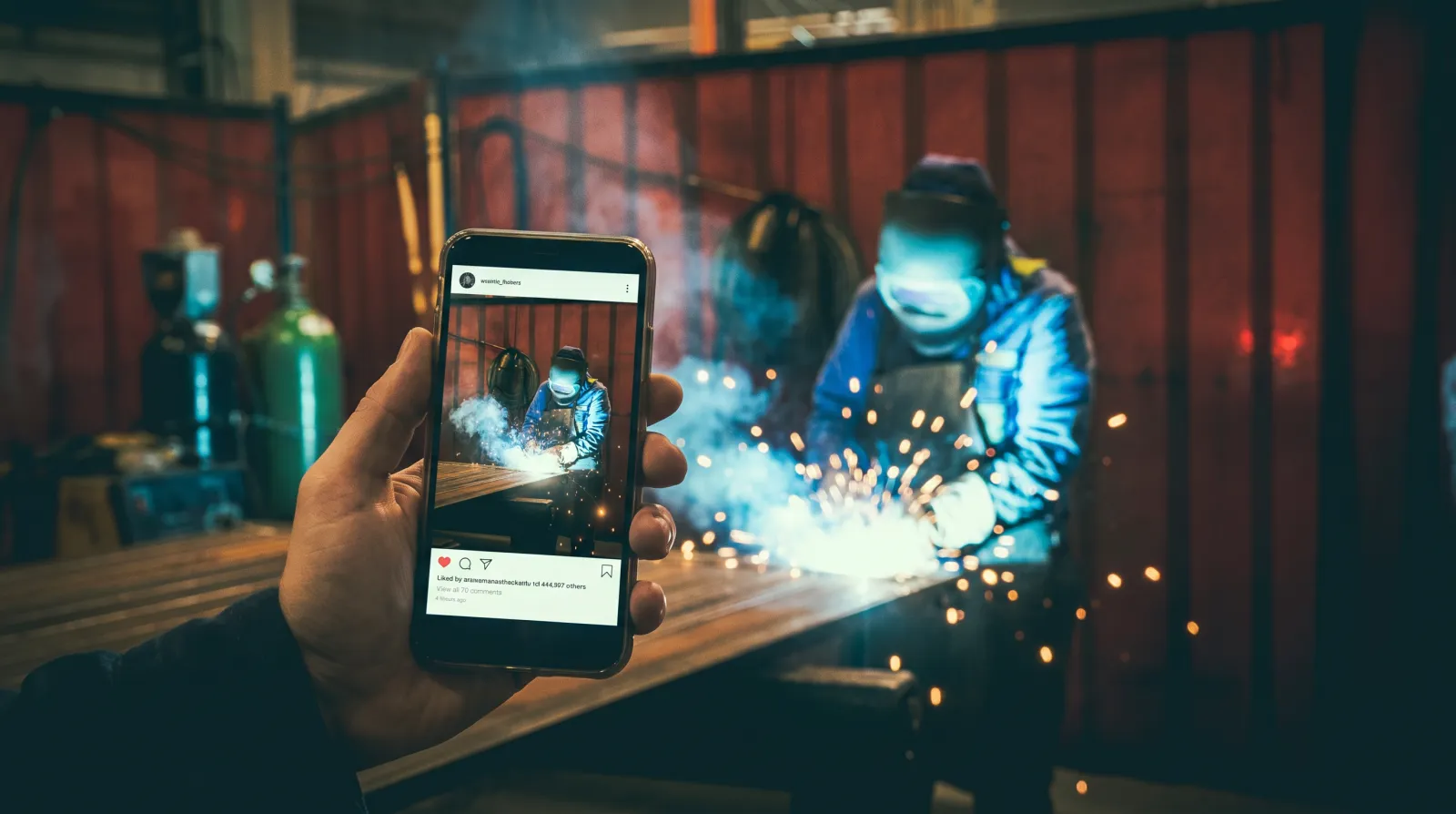 Smartphone mit fertigem Social-Media-Beitrag, im Hintergrund ein Schweißer bei der Arbeit