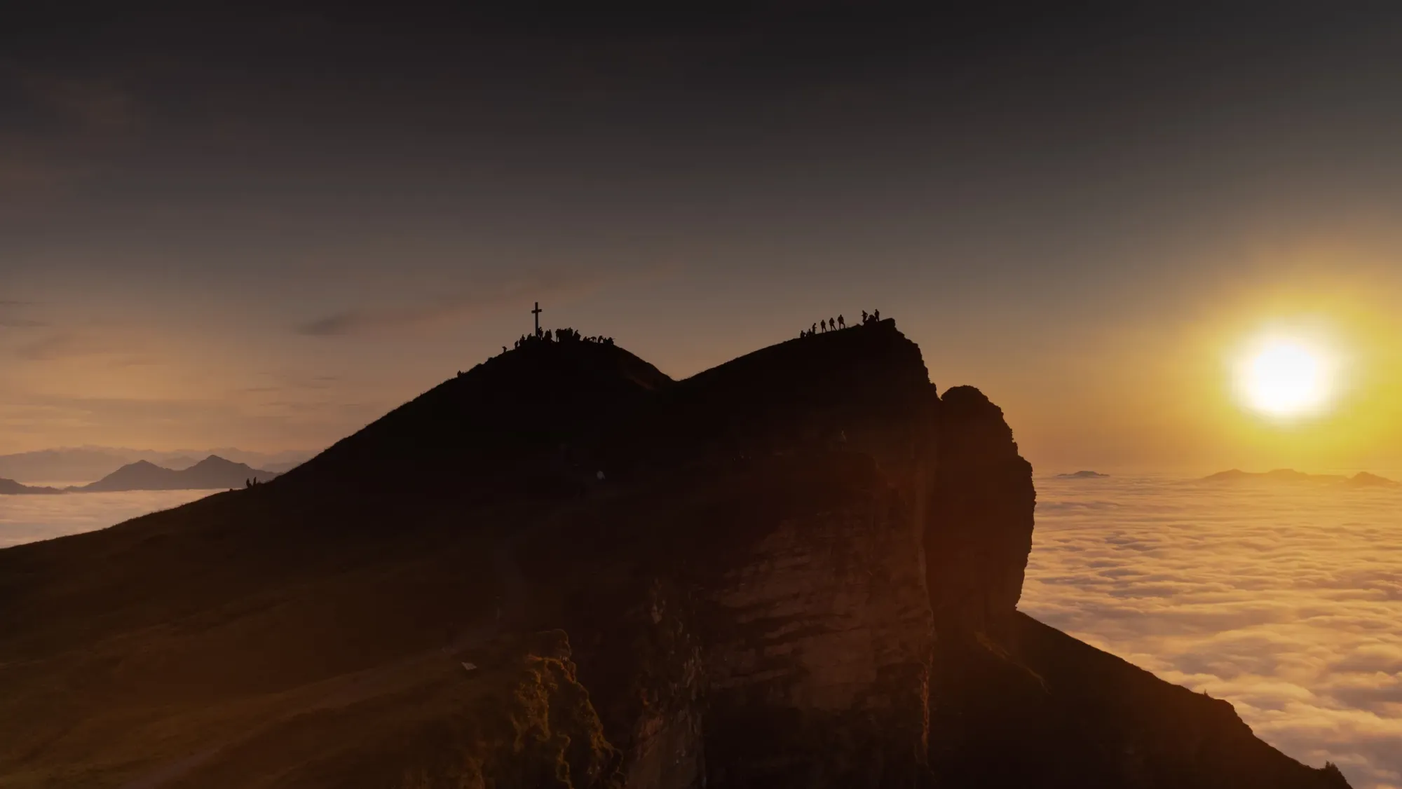 Berggipfel mit Gipfelkreuz über einem Wolkenmeer im warmen Licht des Sonnenuntergangs