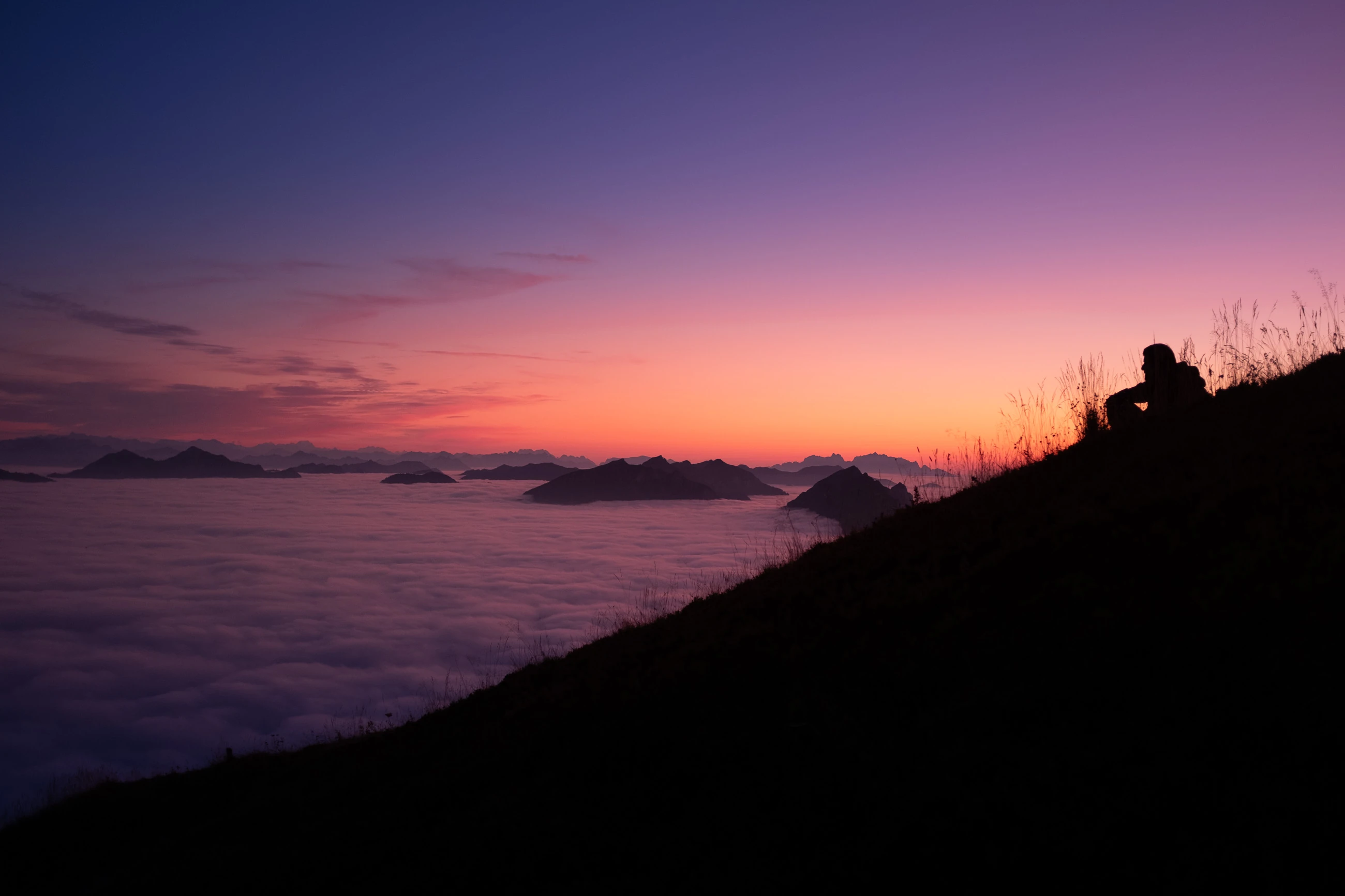 Silhouette einer Person am Berghang mit Blick über ein Nebelmeer und Alpengipfel bei Sonnenuntergang