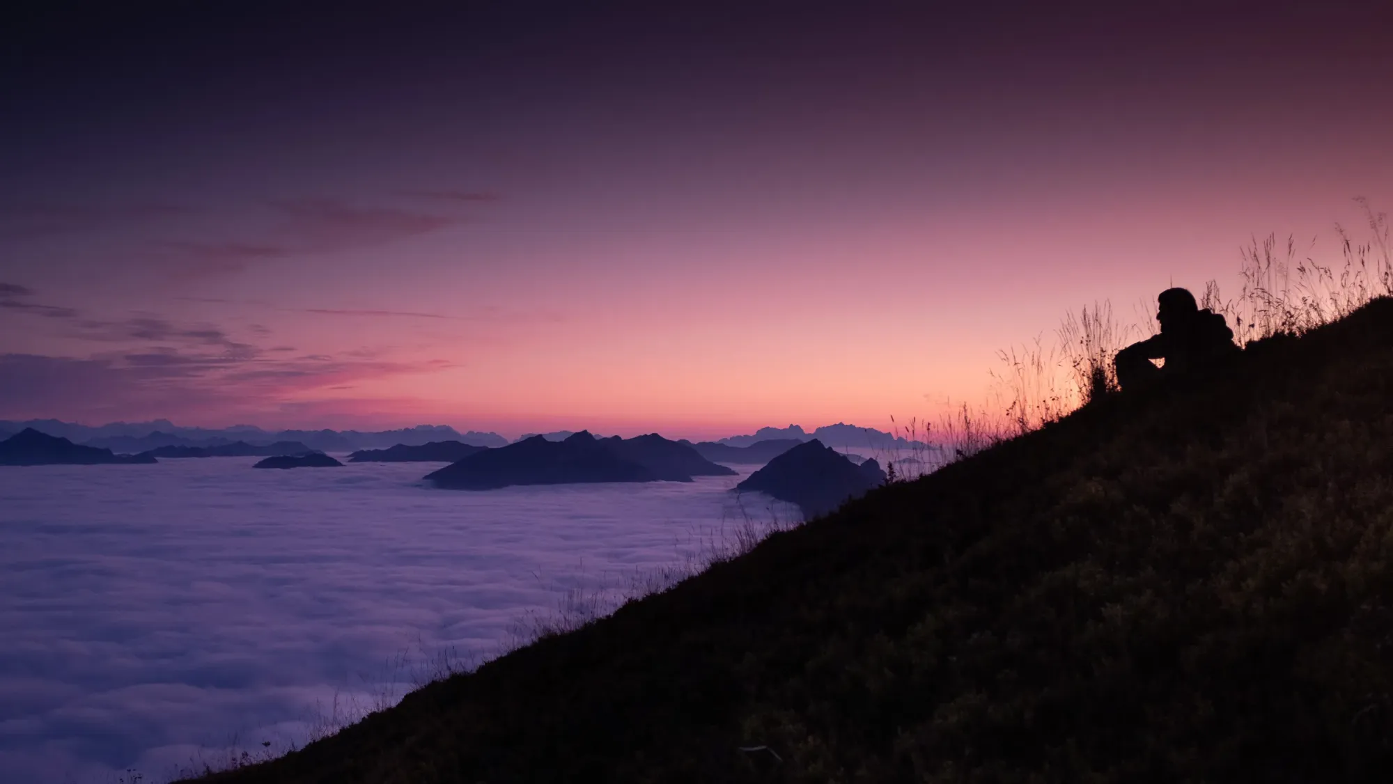 Person am Berghang über einem Wolkenmeer, Alpengipfel in der violetten Dämmerung