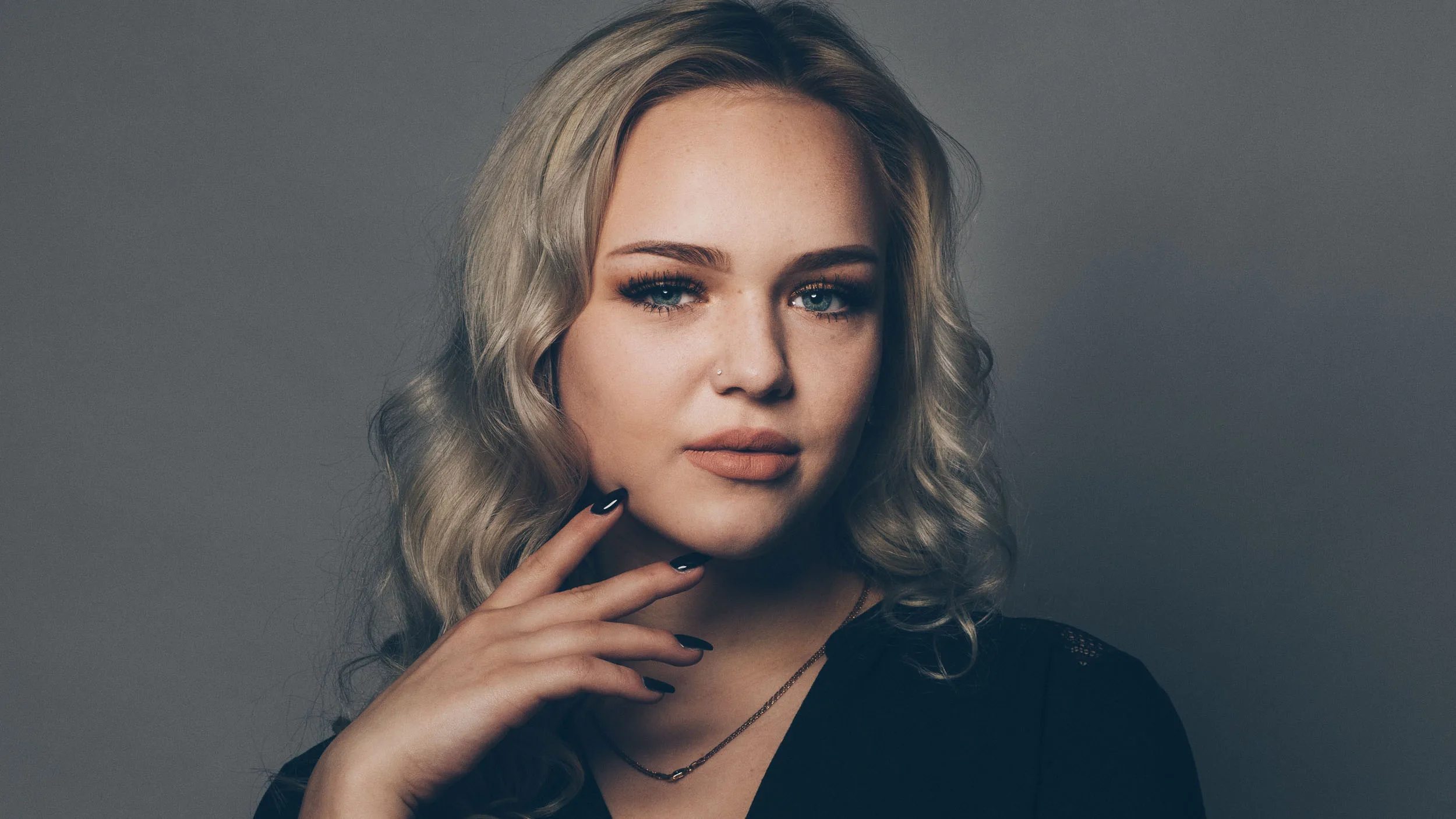 Studio-Porträt einer jungen Frau mit blondem, welligem Haar vor grauem Hintergrund